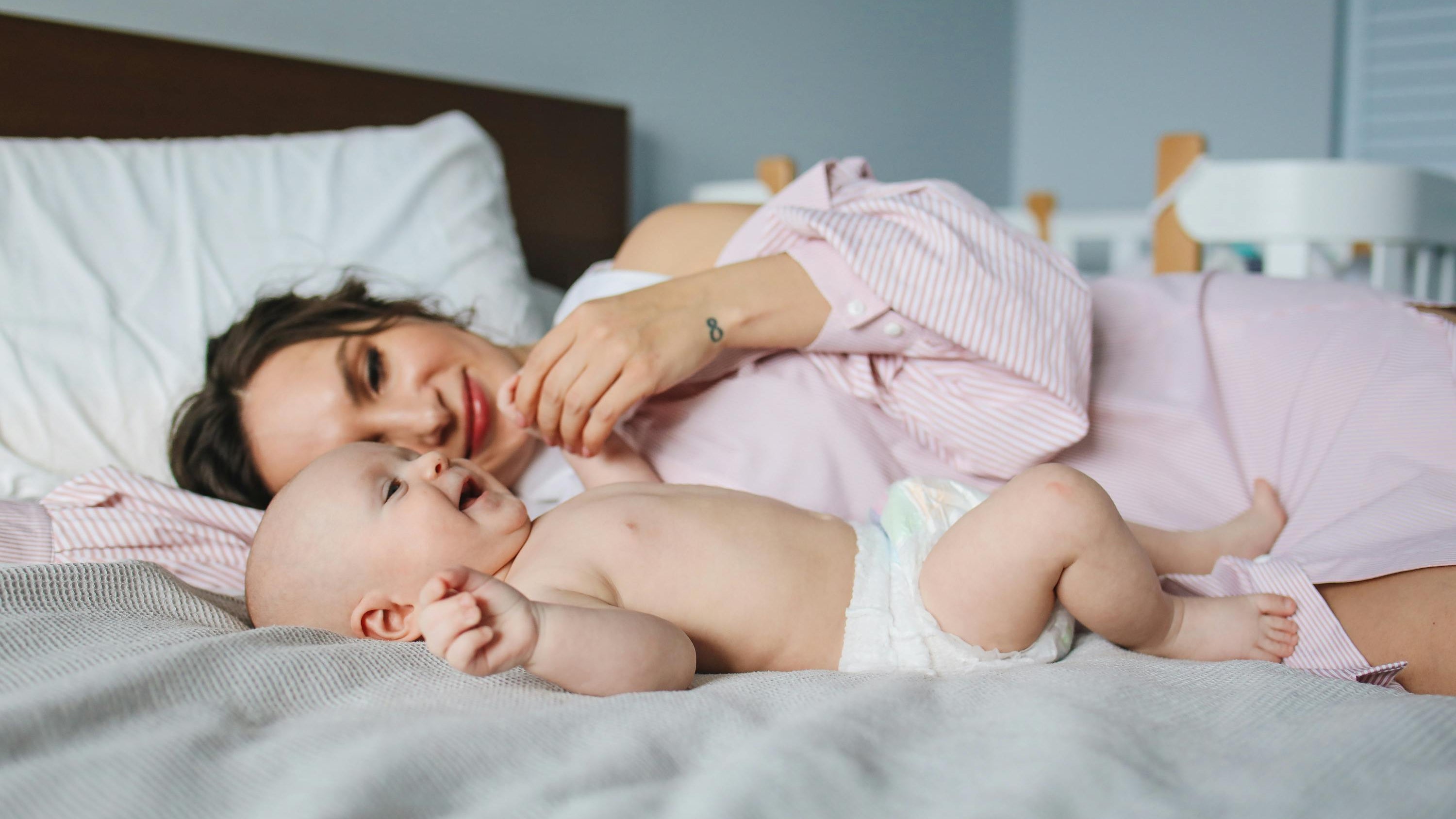 Woman lying on a bed with a baby, both wearing pink outfits.